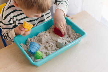 A boy with brown hair is playing at a table with kinetic sand and colorful molds. A boy plays with...