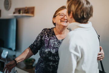 An elderly woman warmly greets another adult in a cozy home environment, showcasing happiness and...
