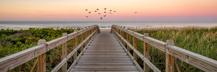Obraz premium Dreamy sunset beach boardwalk, weathered wood path through swaying dune grass, flock of birds soaring under pink-purple sky, serene ocean horizon glow