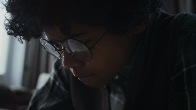 Tilting close-up of hands and face of distressed young African American boy fidgeting with shirt sleeve while reading mocking, humiliating messages from internet trolls on social media on smartphone