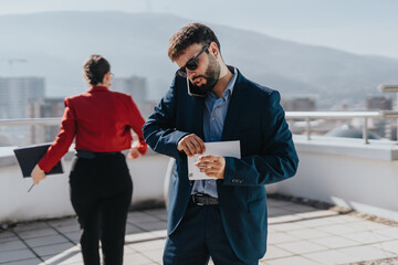 A businessman in formal attire multitasks while on the phone and opening an envelope on a rooftop....