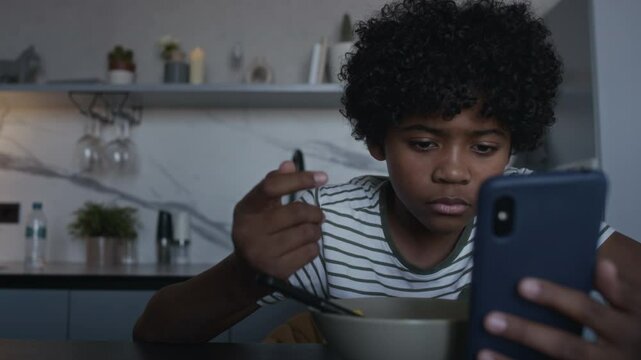 Medium closeup of depressed adolescent African American boy eating cornflakes with milk for breakfast in kitchen, getting vicious message from haters, dropping spoon and typing reply on smartphone