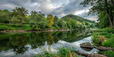 Fototapeta premium Tranquil river mirrors lush forest and golden autumn trees, dramatic clouds above, serene reeds and stone foreground, layered hills in misty depth, cinematic nature dreamscape