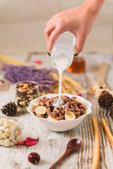 Woman's hand pouring milk from a bottle onto a bowl of chocolate cereal with banana slices on a rustic wooden table, healthy breakfast concept.