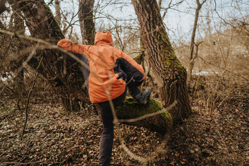 A person in an orange jacket climbs a tree with green moss growing on its bark in a natural, forested environment. The scene emphasizes exploration, nature, and outdoor activities.
