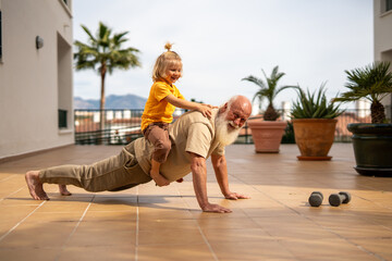 Grandfather doing push-ups while carrying his grandchild on his back, enjoying a fun moment of...