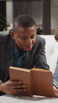 Black private tutor reads book with positive junior schoolboy studying English language at home lesson with native speaker in stylish apartment slow motion