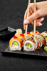 Woman picking up sushi rolls from plate with chopsticks on dark background