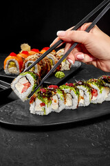 Woman using chopsticks picking sushi rolls from plate on black table