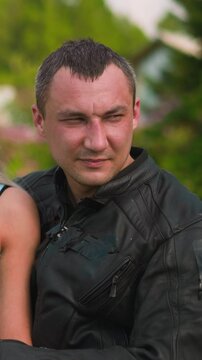 Attractive woman and skilled biker man in leather moto jacket sit on motorcycle against village buildings and gardens on sunny day closeup slow motion