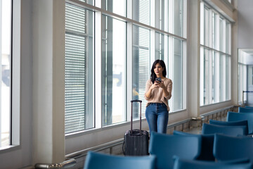 Fototapeta premium Happy Female Travelling use smartphone. Happy asian woman passenger traveler waiting for departure with suitcase at modern airport terminal