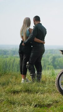 Motorcyclist with slim girlfriend stands near contemporary bike on steep hill against picturesque lake on spring day backside view slow motion