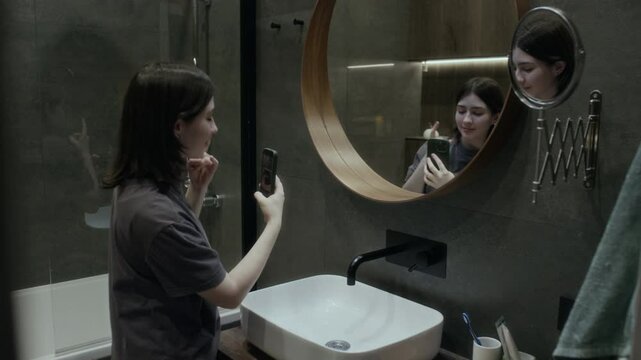 Medium side shot of young teenage Caucasian girl in casual t-shirt standing in bathroom in front of mirror, taking selfies on smartphone for social media, trying different poses and face expressions