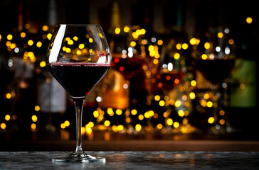 Red merlot wine glass and wine bottles on dark bar background with lights
