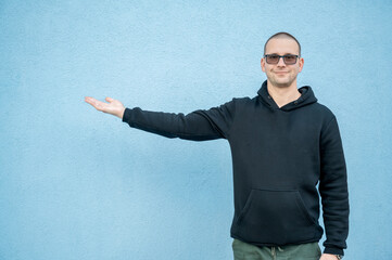 Stylish guy in glasses and black sweatshirt isolated on plain bright blue background, pointing with...