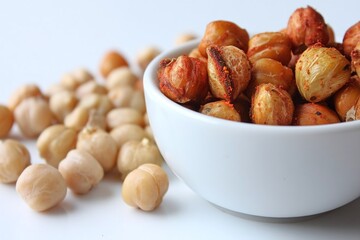 Obraz premium Bowl of roasted nuts with scattered raw nuts on a white surface, showcasing a healthy snack option