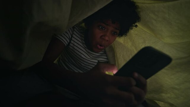 Medium shot of cheerful, excited 13-year-old African American boy lying in bed at night, hiding under duvet, viewing inappropriate or entertaining content on smartphone with surprise