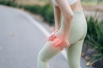 woman in activewear is outdoors on pathway, holding her thigh with pained expression, indicating muscle discomfort or injury. scene suggests physical strain during exercise