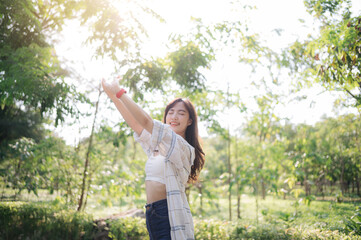 Fototapeta premium Asian woman enjoying summer while breathing fresh air in lush green park, arms outstretched, feeling relaxed and happy under sunlight