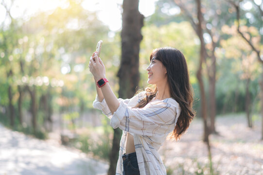 cheerful young woman taking selfie in sunlit park, wearing casual plaid shirt and smartwatch, surrounded by lush greenery and soft sunlight