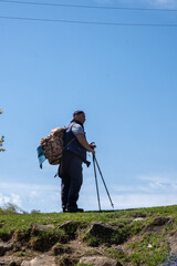 Vertical shot: An overweight adult male hiker wearing a cap and carrying a backpack leans on trekking poles, gazing into the distance, anticipating a solo hiking adventure.