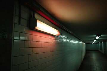 Orange emergency light flickering and illuminating a dark subway corridor, revealing tiled walls and casting eerie shadows in the underground tunnel