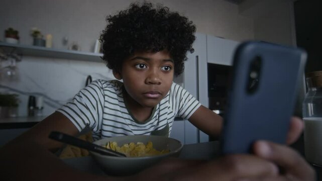 Medium shot of 13-year-old African American boy eating cornflakes in morning in kitchen, getting message with mature or inappropriate content, checking around, viewing it and grinning