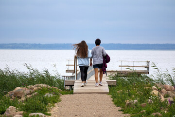 
Two people, a man and a woman, walking from behind along a wooden pier towards a calm body of water under a cloudy sky.