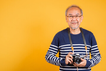 Portrait Asian old man wearing glasses holding vintage SLR camera studio shot isolated yellow background, smiling happy Photographer elderly man gray haired holds old fashioned camera