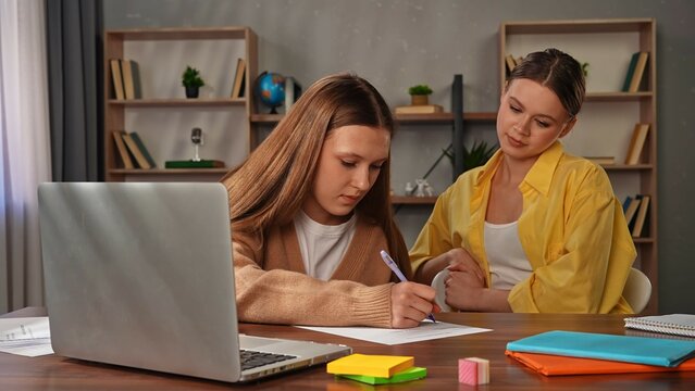Young teenage girl studying at home, girl with mother sit at desk with laptop and books and doing homework test, circling answers on paper, mom controls process.