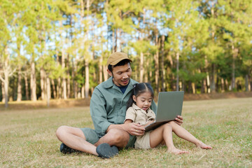 Obraz premium Asian male father and young Asian girl sitting together on grassy field using laptop, smiling and interacting happily outdoors, expressing strong family bond, education, and love of learning nature.