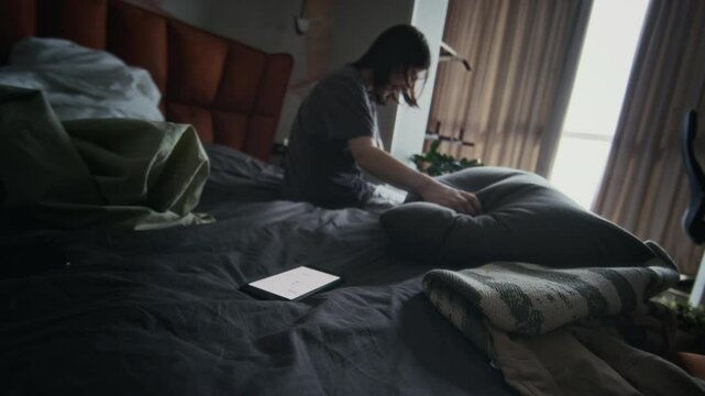 Wide rolling zoom-in shot of desperate bullied teenage girl sitting on unkempt bed at home, weeping, thumping and pounding on pillow in rage and anger. Smartphone with abusive messages nearby