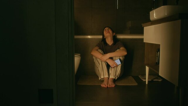 Wide shot of heartbroken young Caucasian girl in pyjamas crouching on floor in bathroom, clutching smartphone, sobbing and weeping, due to betrayal or online harassment