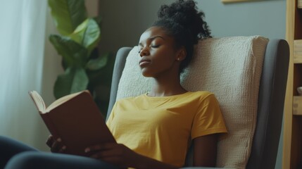 Sideways young woman of African American ethnicity wear t-shirt drink coffee read book close eyes sits in armchair stay at home flat rest relax spend free spare time in living room indoor grey wall.