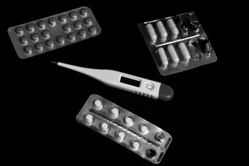 Black and white horizontal photo, various pills in blister packs and a thermometer on a black background