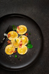 fresh ravioli, with butter, spices and herbs, fried bacon, on a black plate, top view, dark background, no people,