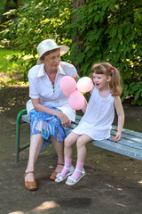Grandmother and granddaughter with balloons shares smile, sitting on a bench in a garden. Happy family moment with two people, an elderly woman and a young baby girl, enjoying time together outdoors