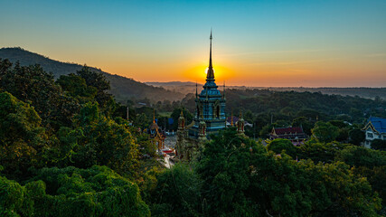 Fototapeta premium A breathtaking aerial view of a golden Buddhist temple at Wat Mae Klang Waterfall in Chiang Mai Thailand is surrounded by lush greenery, with the sunrise casting warm hues across the horizon.