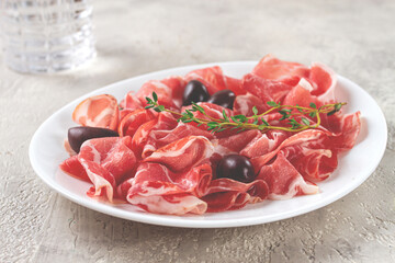 Freshly cut dried meat, served with olives and herbs, on a white plate, light background, no people,