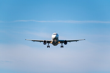 Front view of a commercial airplane approaching for landing against a bright blue sky with scattered clouds.