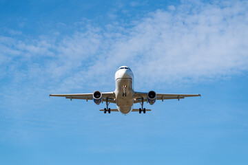 Front view of a commercial airplane approaching for landing against a bright blue sky with scattered clouds.
