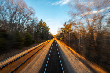 Motion blur view looking out the back of a moving passenger train speeding through a forested railway at sunset, with clear skies above.