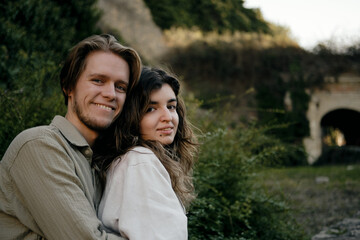Close-up portrait of a young smiling couple embracing outdoors, surrounded by lush greenery. Natural and authentic love story moment. Woman and man in love on date in park.