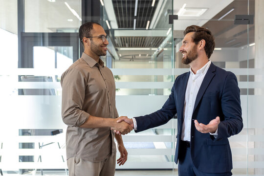 Two businessmen shake hands in a modern office, smiling warmly during a professional agreement or greeting, with natural light.