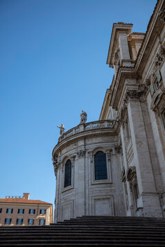 Low angle view of the apse and grand steps of the Basilica di Santa Maria Maggiore featuring statues and classical architectural details against a blue sky in Rome, Lazio, Italy on a sunny day.