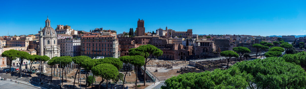 Panoramic view of the Imperial Fora, Trajan's Column, and Santa Maria di Loreto church with pine trees under a clear blue sky in Rome, Italy, ancient Roman ruins cityscape landscape architecture.
