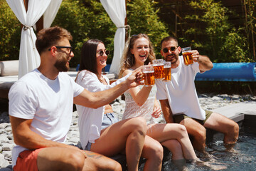Group of cheerful friends toasting with beer while relaxing by the pool on a sunny summer day