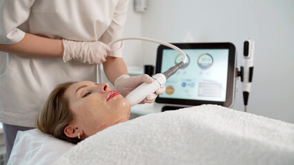 Obraz premium Beautician using special equipment performing red light therapy treatment on woman's face in beauty salon