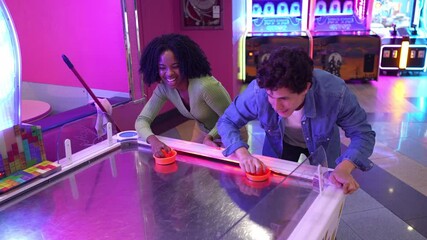 Energetic multi ethnic couple competing and laughing while playing air hockey, sharing playful moments inside colorful arcade gaming environment