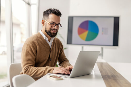 Young smiling data analyst sitting at corporate office and typing on a laptop.